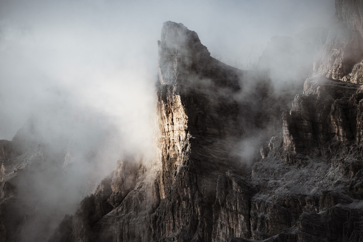 A rugged mountain peak gets illuminated by a sunray surrounded by fog